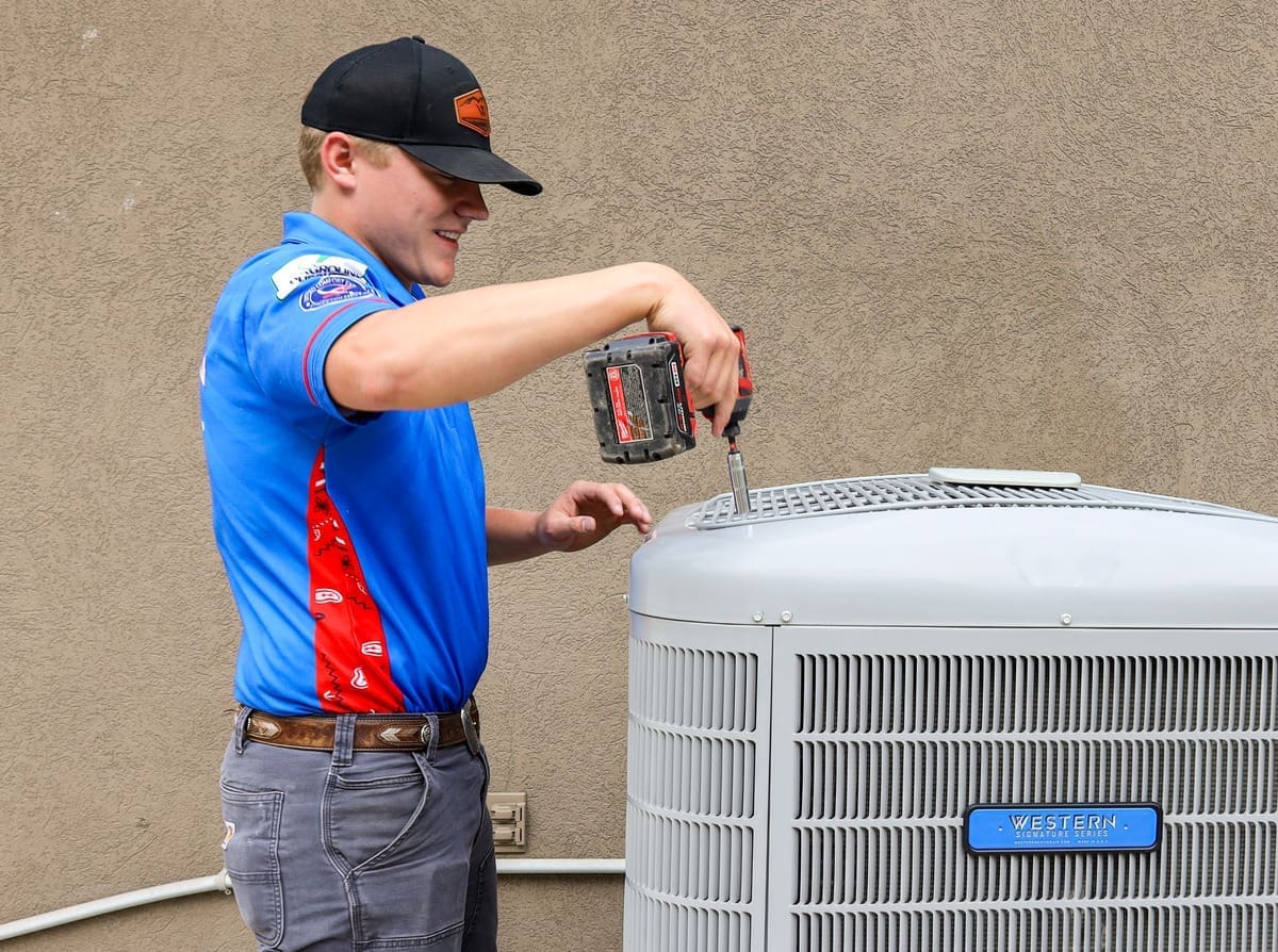 This image shows a professional performing AC installation in Lehi UT at a residential property. The technician from Western Heating, Air & Plumbing is securing the top panel of an outdoor air conditioning condenser using a drill while standing beside the unit. The setup reflects a clean and organized work area with visible HVAC components and proper installation steps. Western Heating, Air & Plumbing serves homeowners across Utah County with reliable air conditioning installation, helping families maintain indoor comfort with quality workmanship and trusted service.