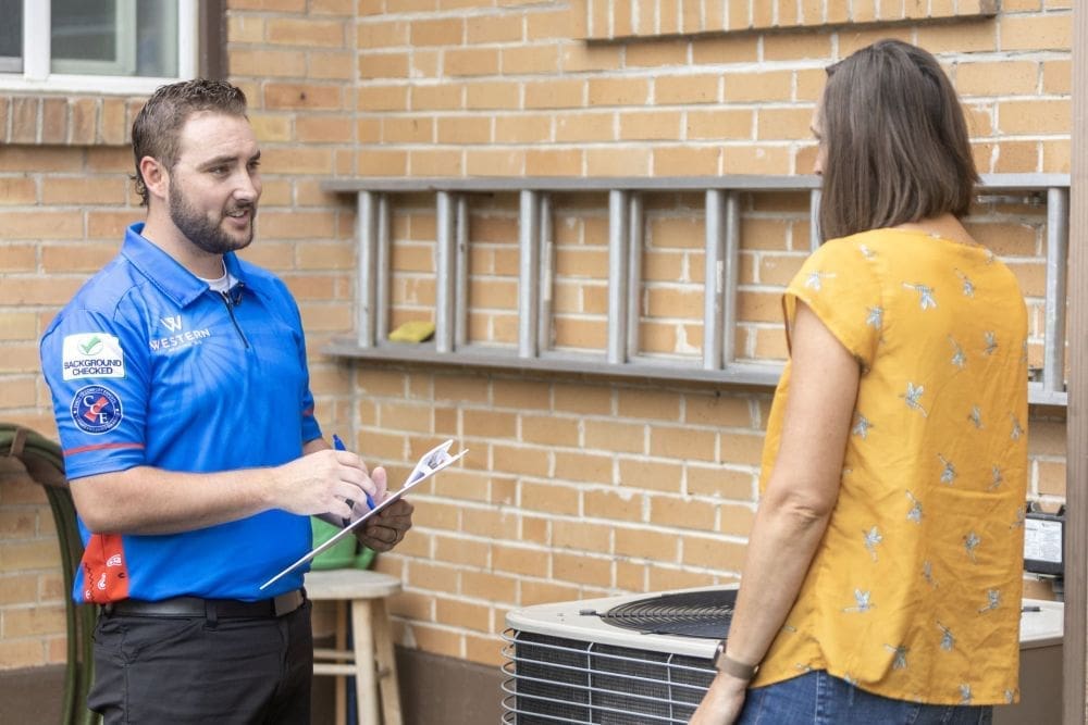 AC replacement in Orem UT consultation with a homeowner and HVAC technician standing beside an outdoor condenser unit. Western Heating, Air & Plumbing provides AC replacement services in Orem, Utah, helping homeowners understand system options and upgrade aging air conditioning units. The image shows a technician explaining replacement details while holding a clipboard, with the homeowner listening near the existing AC system. Residents in Utah County trust Western Heating, Air & Plumbing for air conditioner replacement, system upgrades, and reliable cooling solutions. Contact Western Heating, Air & Plumbing in Orem UT for AC replacement consultations and installation services.